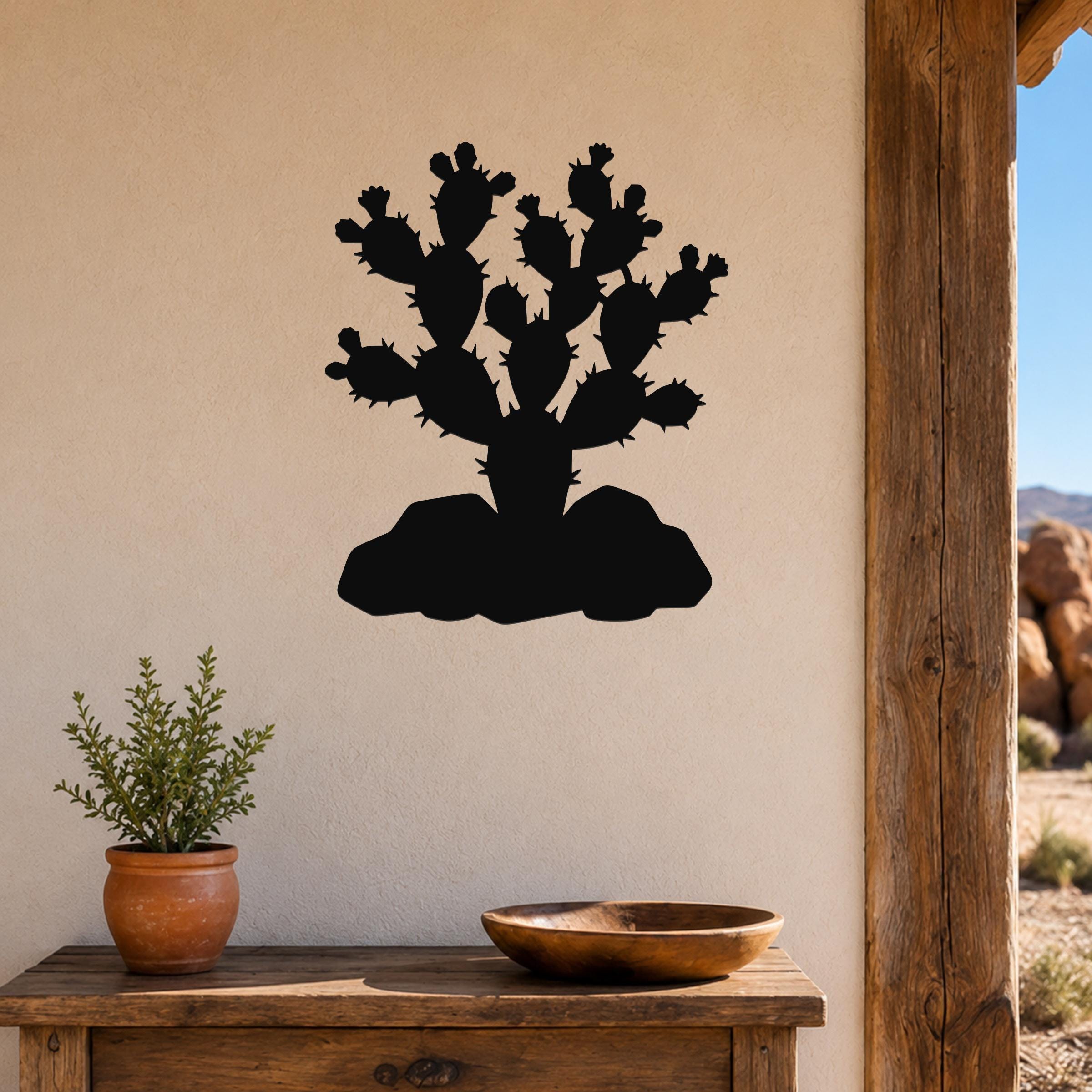 Black metal prickly pear cactus wall art with spiny pads and base silhouette, mounted on a beige wall above a wooden table in a rustic indoor setting.