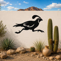 Black metal quail wall art in a running silhouette, mounted on a white outdoor wall above a desert garden with cacti and rocks.