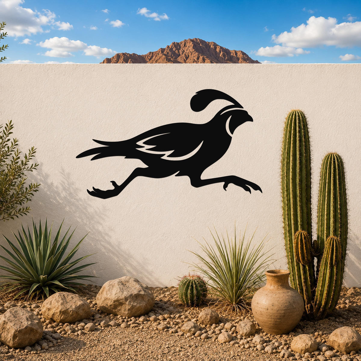 Black metal quail wall art in a running silhouette, mounted on a white outdoor wall above a desert garden with cacti and rocks.