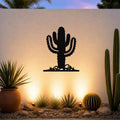 Black metal Saguaro cactus wall art with spines and base cutouts, mounted on a textured outdoor wall above a desert garden with potted plants and ground lights.
