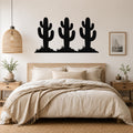 Black metal wall art featuring a silhouette of three saguaro cacti with spines and small plants, mounted above a wooden bed in a bedroom.