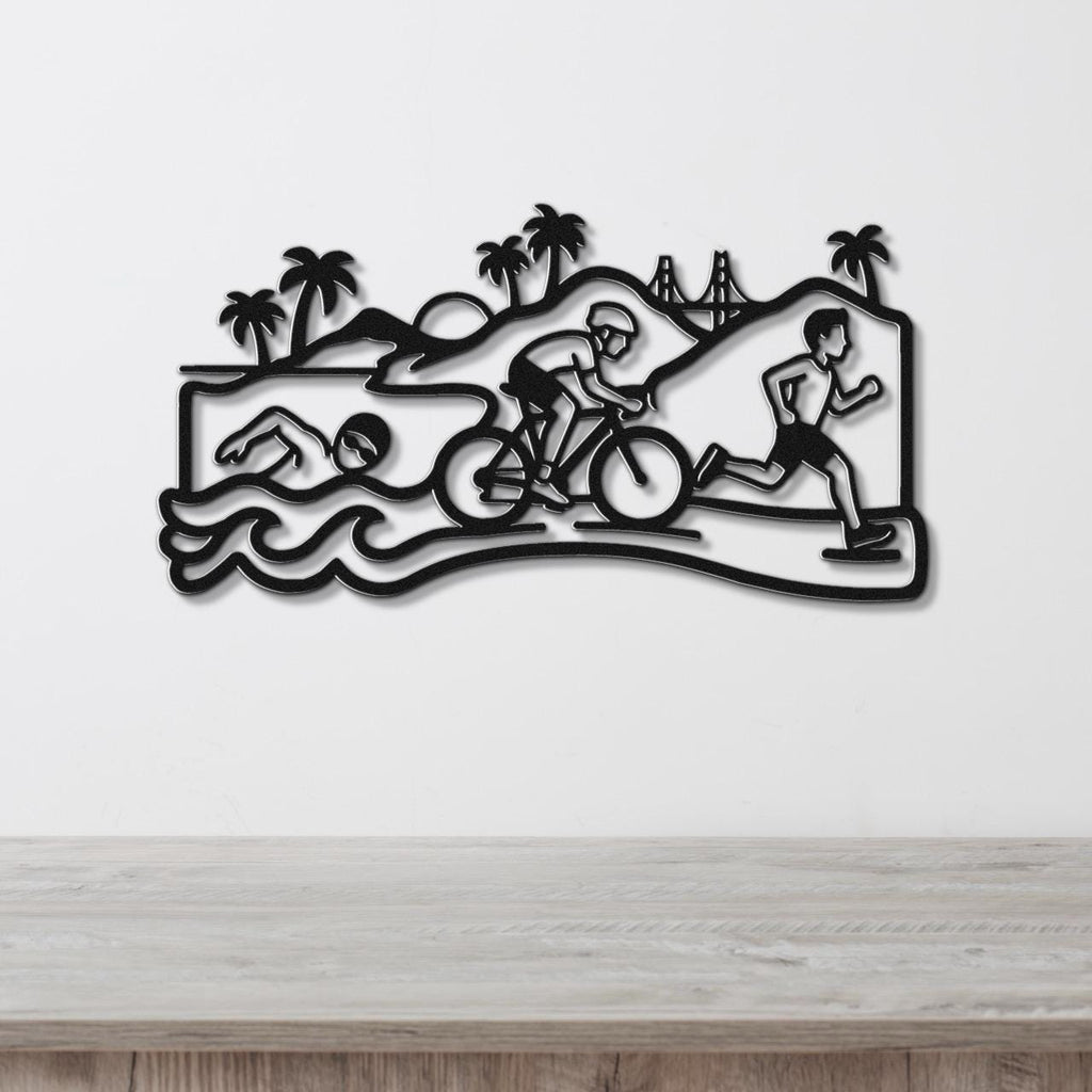 Black metal wall art featuring a triathlon scene with swim, bike, and run silhouettes, palm trees, and a bridge, mounted on a white wall above a wooden surface.