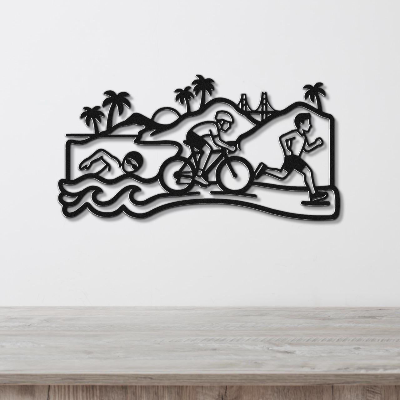 Black metal wall art featuring a triathlon scene with swim, bike, and run silhouettes, palm trees, and a bridge, mounted on a white wall above a wooden surface.