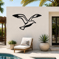 Black metal seagull wall art with wing and tail cutouts, mounted on a white outdoor wall above a lounge chair near a pool.