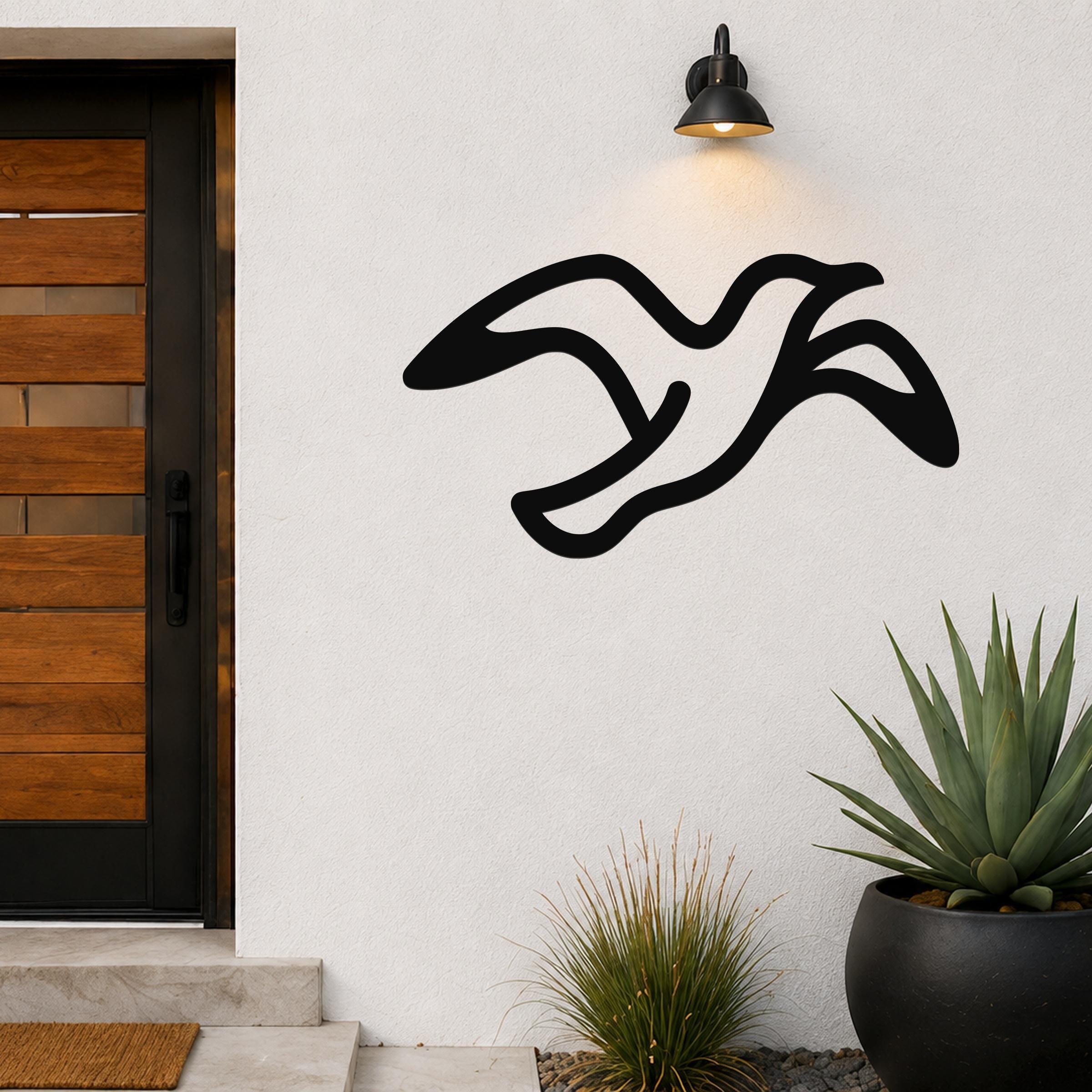 Black metal seagull wall art with flowing motion lines, mounted on a white exterior wall above a potted succulent and near a wooden front door.