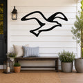 Black metal seagull wall art with flowing wing cutouts, mounted on a white shiplap wall above a wooden bench on a porch.