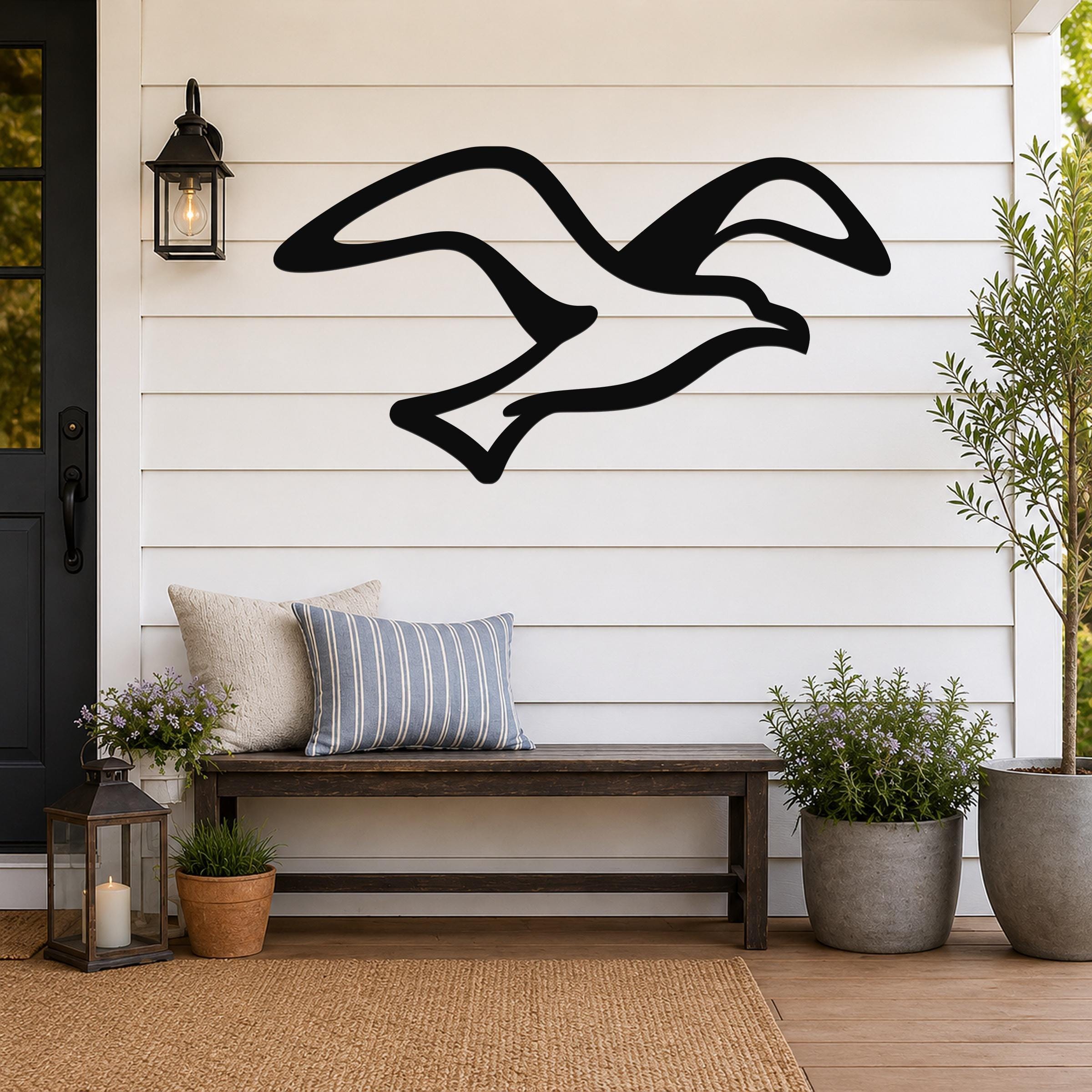 Black metal seagull wall art with flowing wing cutouts, mounted on a white shiplap wall above a wooden bench on a porch.