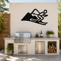Black metal skier and mountain wall art silhouette mounted above a stainless steel outdoor grill on a patio wall.