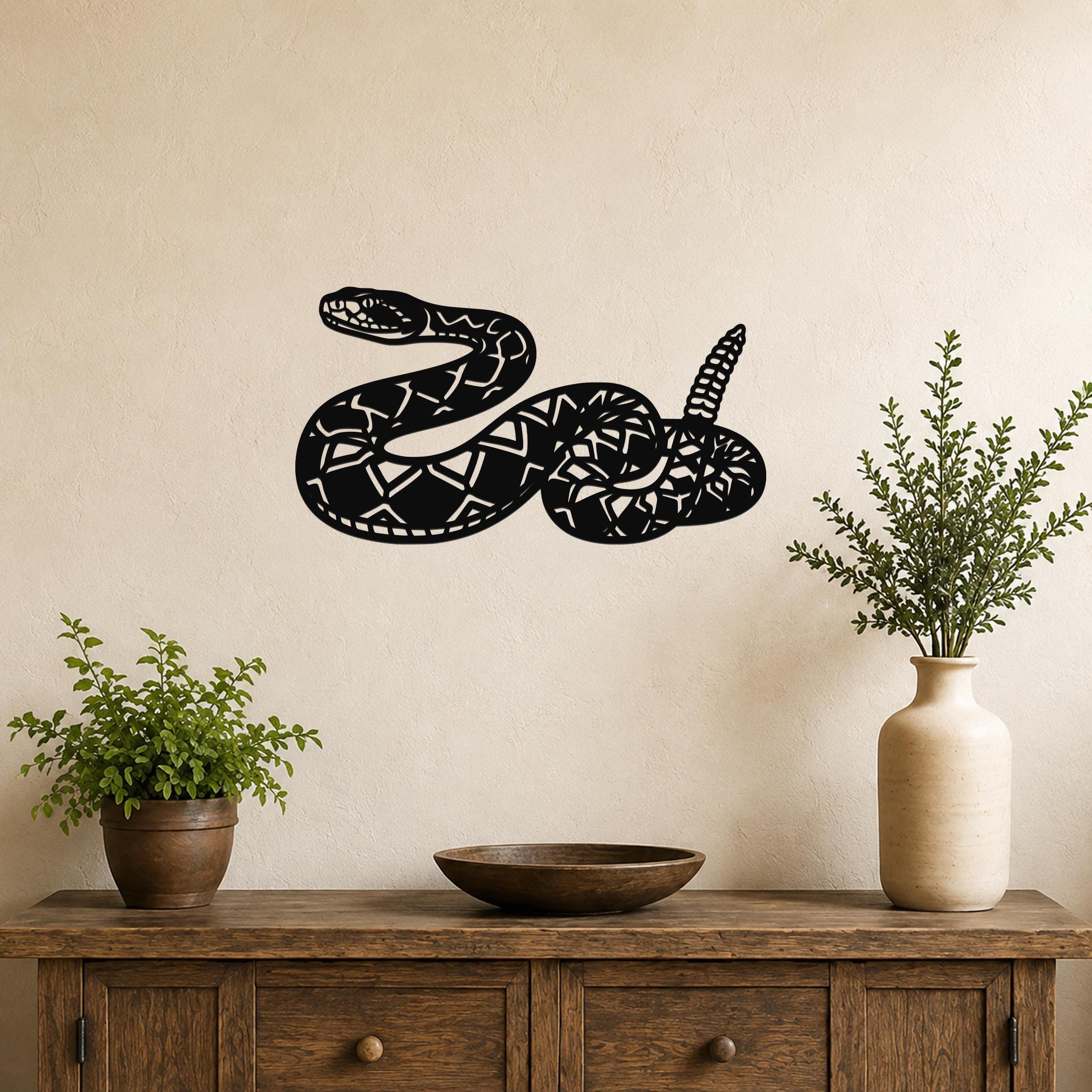 Black metal wall art sculpture of a coiled rattlesnake with detailed scale and rattle cutouts, mounted on a beige wall above a wooden console table with plants.