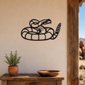 Black metal wall art of a coiled rattlesnake silhouette with detailed internal cutouts, mounted on a beige wall above a wooden table in a rustic outdoor patio setting.