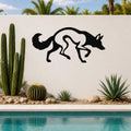 Black metal coyote wall art with a silhouette cutout design, mounted on an outdoor wall above a pool with cacti and palm trees in the background.