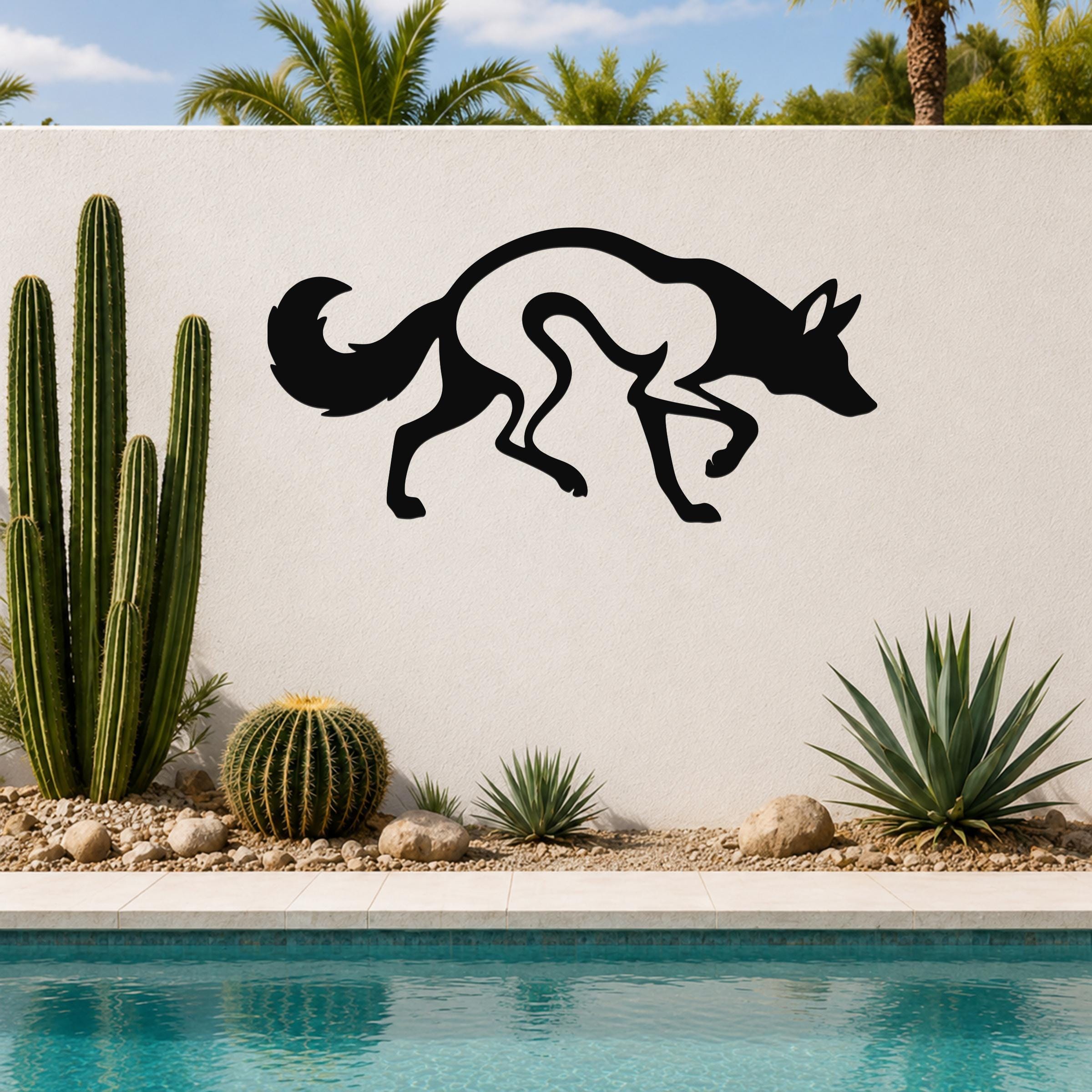 Black metal coyote wall art with a silhouette cutout design, mounted on an outdoor wall above a pool with cacti and palm trees in the background.