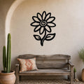 Black metal sunflower wall art with a spiral center and leaf cutouts, mounted on a textured beige wall above a rustic wooden bench in a patio setting.