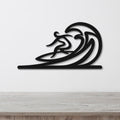 Black metal wall art featuring a surfer silhouette riding a flowing wave, mounted on a white wall above a wooden surface.