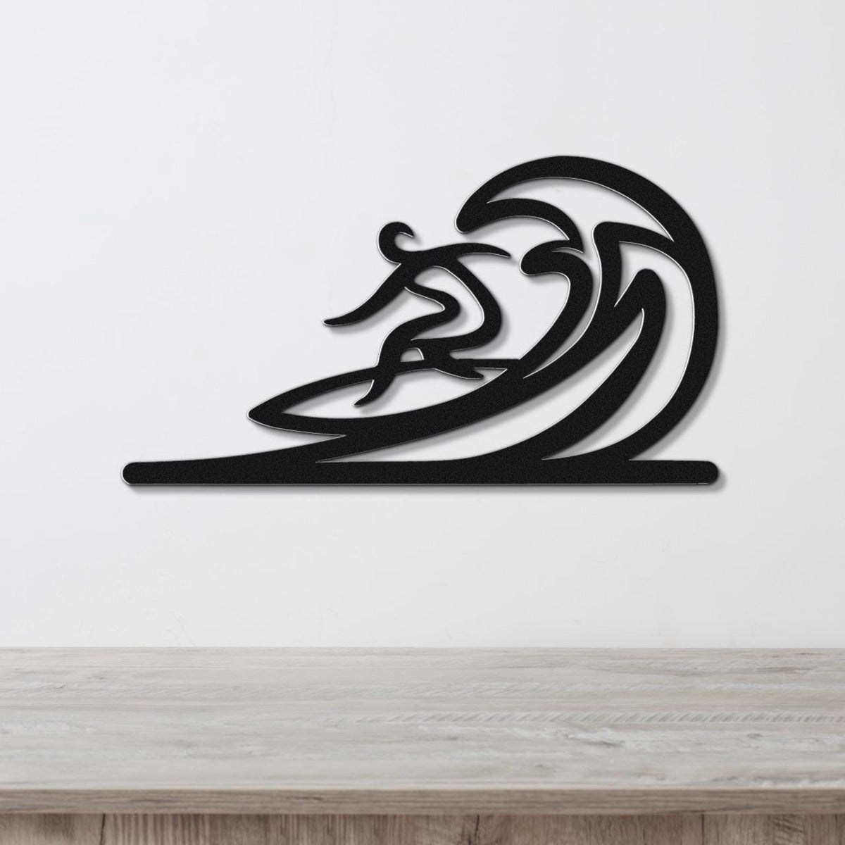 Black metal wall art featuring a surfer silhouette riding a flowing wave, mounted on a white wall above a wooden surface.