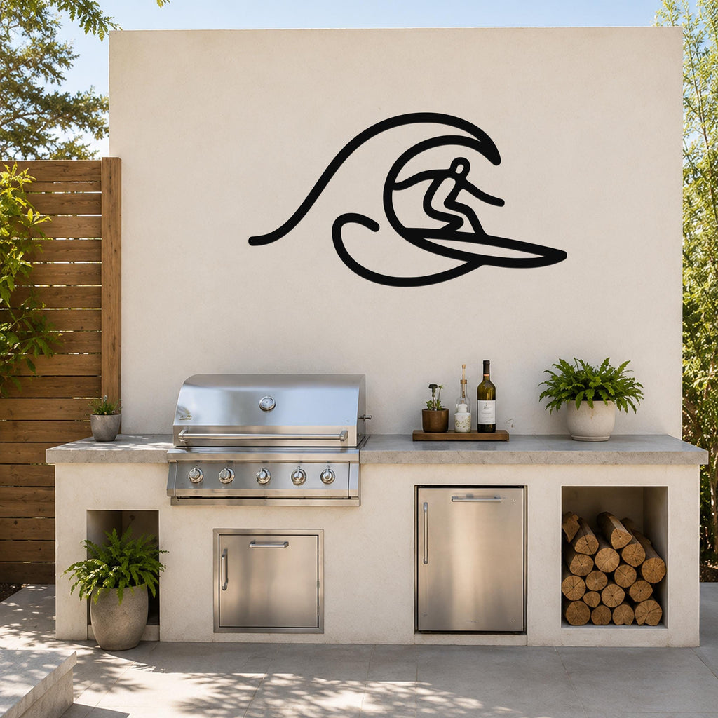 Black metal wall art silhouette of a surfer riding a wave, mounted on an outdoor kitchen wall above a stainless steel grill and counter.