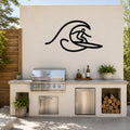 Black metal wall art silhouette of a surfer riding a wave, mounted on an outdoor kitchen wall above a stainless steel grill and counter.