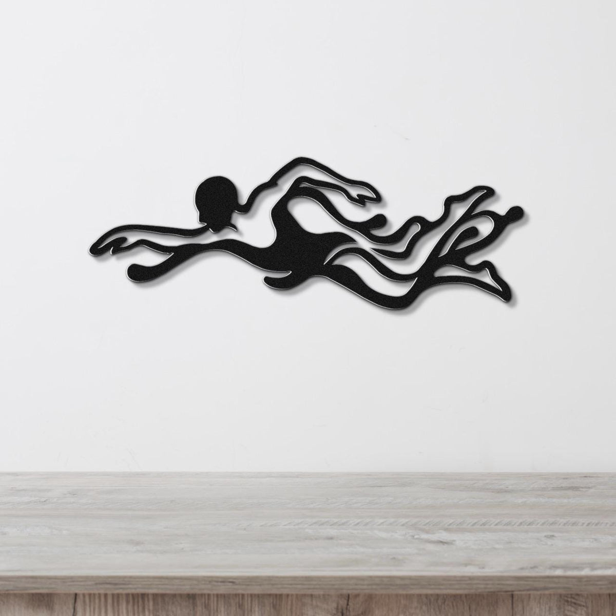Black metal wall art of a swimmer in motion with flowing water cutouts, mounted on a white wall above a light wood surface.