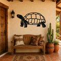 Black metal tortoise wall art with detailed shell and limb cutouts, mounted on a stucco wall above a wooden bench in a patio entryway.