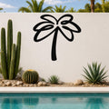 Black metal palm tree wall art with flowing frond cutouts, mounted on a white outdoor wall above a pool with cacti and succulents.