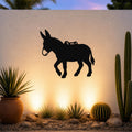 Black metal burro wall art silhouette with saddle cutout, mounted on an outdoor wall above a desert garden with cacti and warm uplighting.
