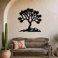 Black metal wall art silhouette of a Joshua tree with spiky branches and rocky base, mounted on a textured beige wall above a wooden bench in a southwestern-style living space.