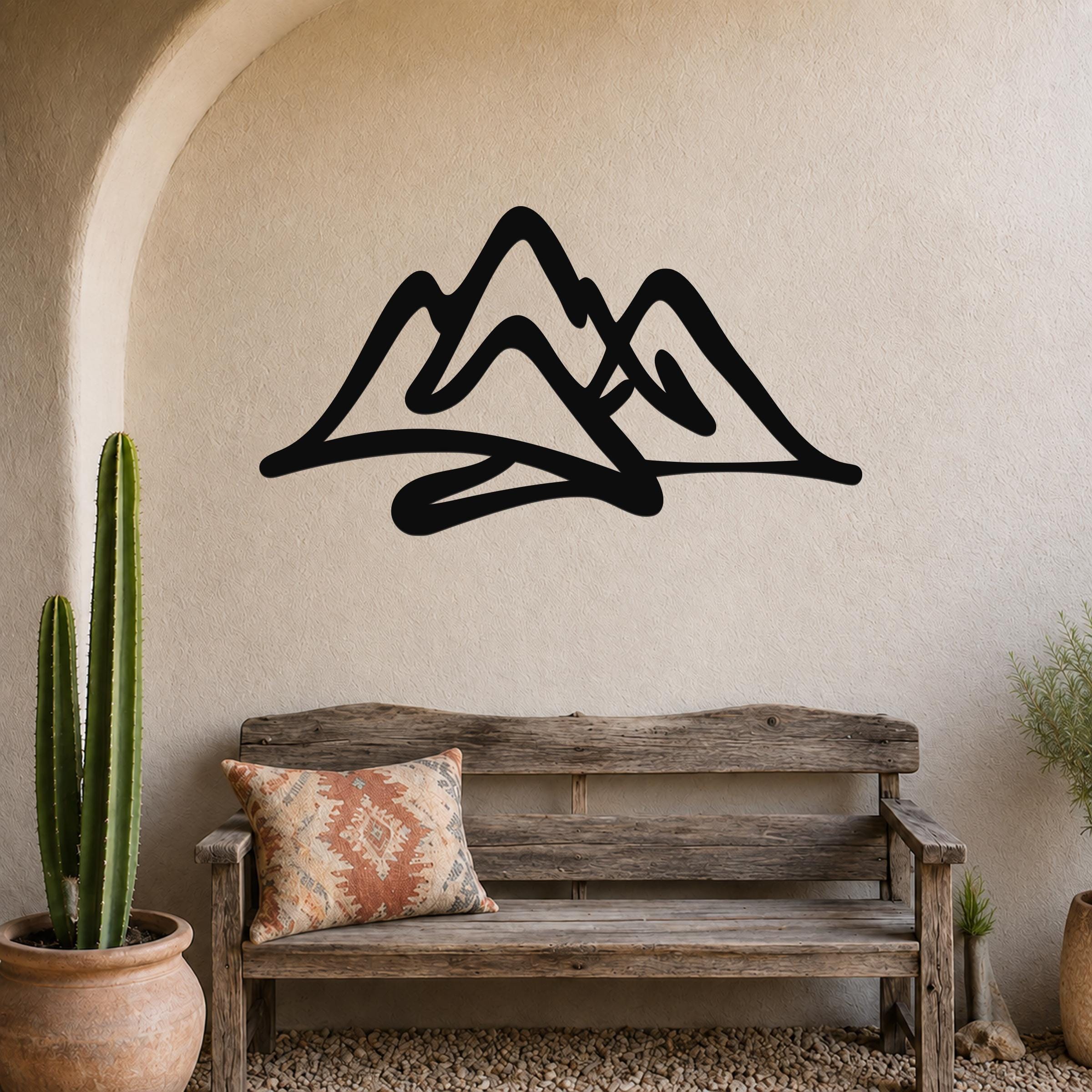 Black metal mountain silhouette wall art mounted above a rustic wooden bench in a desert-style room with cacti and pebbles.