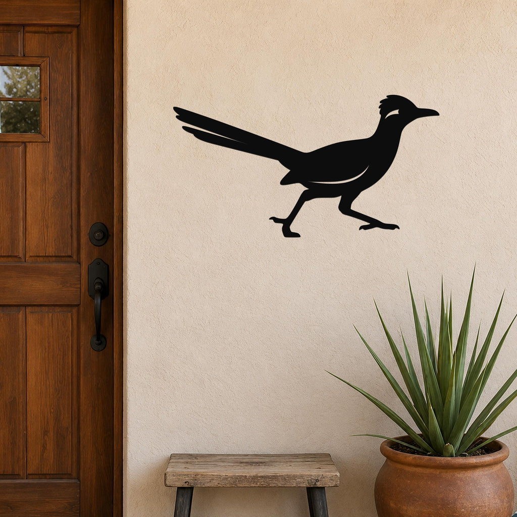 Black metal roadrunner wall sculpture with long tail and crest, mounted on a beige stucco wall beside a wooden door and potted plant.