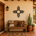 Black metal Zia Sun symbol wall art with bold circular design and radiating lines, mounted on a stucco wall above a wooden bench in a Southwestern-style patio.