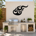Black metal flame wall art with flowing silhouette design, mounted on an outdoor kitchen wall above a stainless steel grill and counter.
