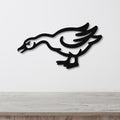 Black metal wall art cutout of a goose in motion, mounted on a white wall above a light wood surface.