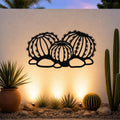 Black metal barrel cactus wall art with detailed spines and segmented cutouts, mounted on a white outdoor wall above a desert garden with potted plants and ground lights.