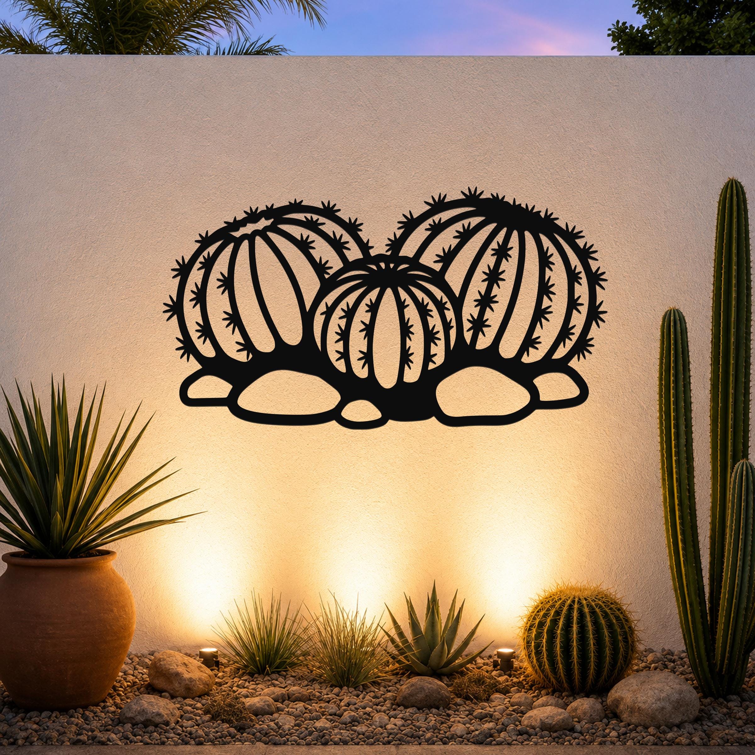Black metal barrel cactus wall art with detailed spines and segmented cutouts, mounted on a white outdoor wall above a desert garden with potted plants and ground lights.