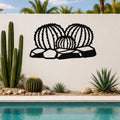 Black metal barrel cactus wall art with ribbed cutout design, mounted on a white outdoor wall above a poolside garden.