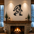 Black metal Bigfoot wall art silhouette mounted above a stone fireplace in a rustic living room with wooden beams and snow-covered trees visible through windows.