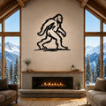 Black metal Bigfoot silhouette wall art mounted above a fireplace in a rustic living room with large windows overlooking snowy mountains.