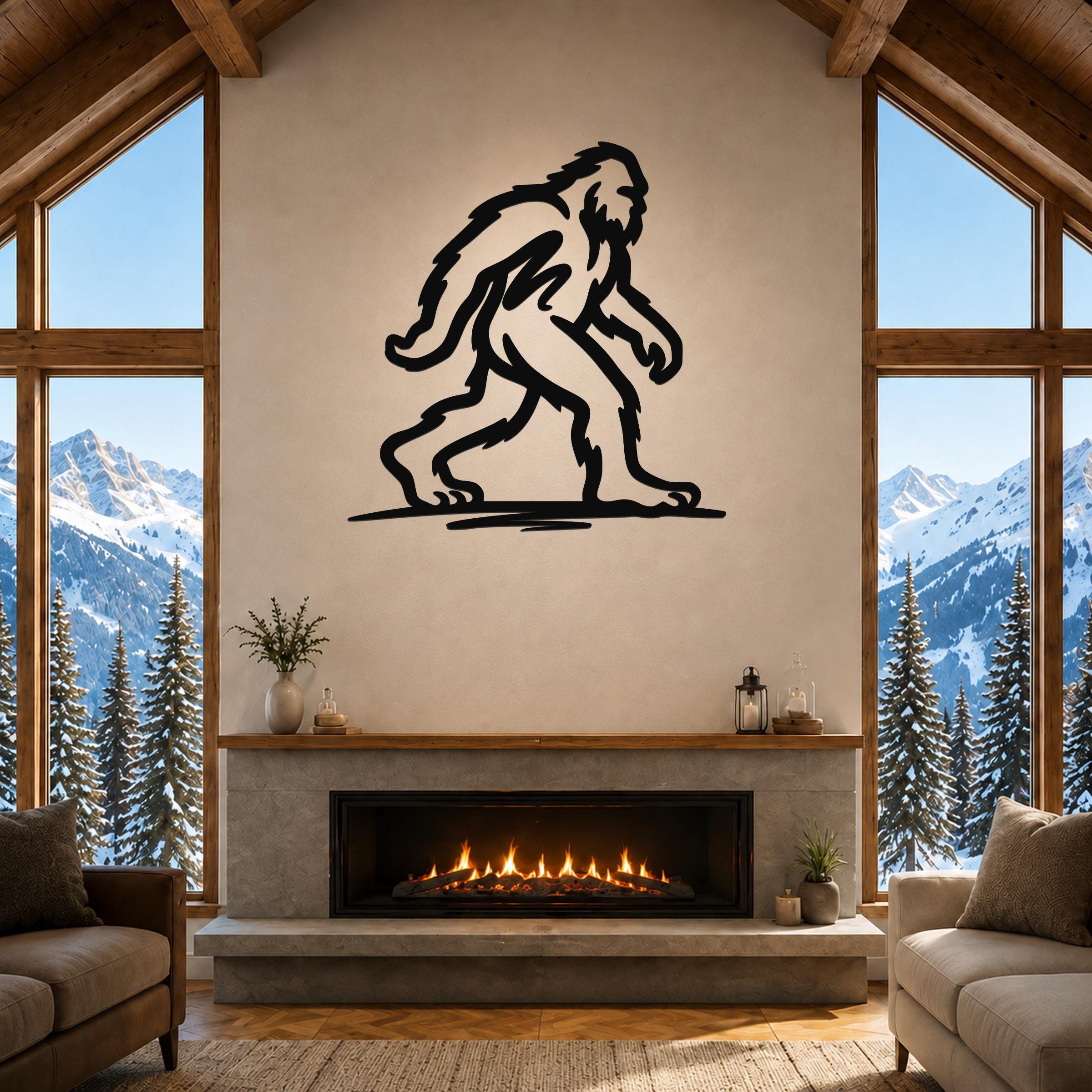 Black metal Bigfoot silhouette wall art mounted above a fireplace in a rustic living room with large windows overlooking snowy mountains.