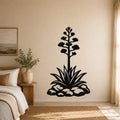 Black metal agave wall sculpture with blooming flower and leaf cutouts, mounted on a beige bedroom wall above a bed.
