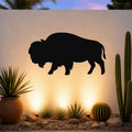 Black metal bison silhouette wall art mounted on a light-colored outdoor wall above a desert garden with cacti and ground lights.