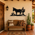 Black metal wall art silhouette of a burro and cacti, mounted on a stucco wall above a wooden bench in a rustic patio entry.
