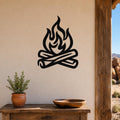 Black metal campfire wall art with flame and log silhouette, mounted on a beige wall above a wooden table in a rustic outdoor patio setting.