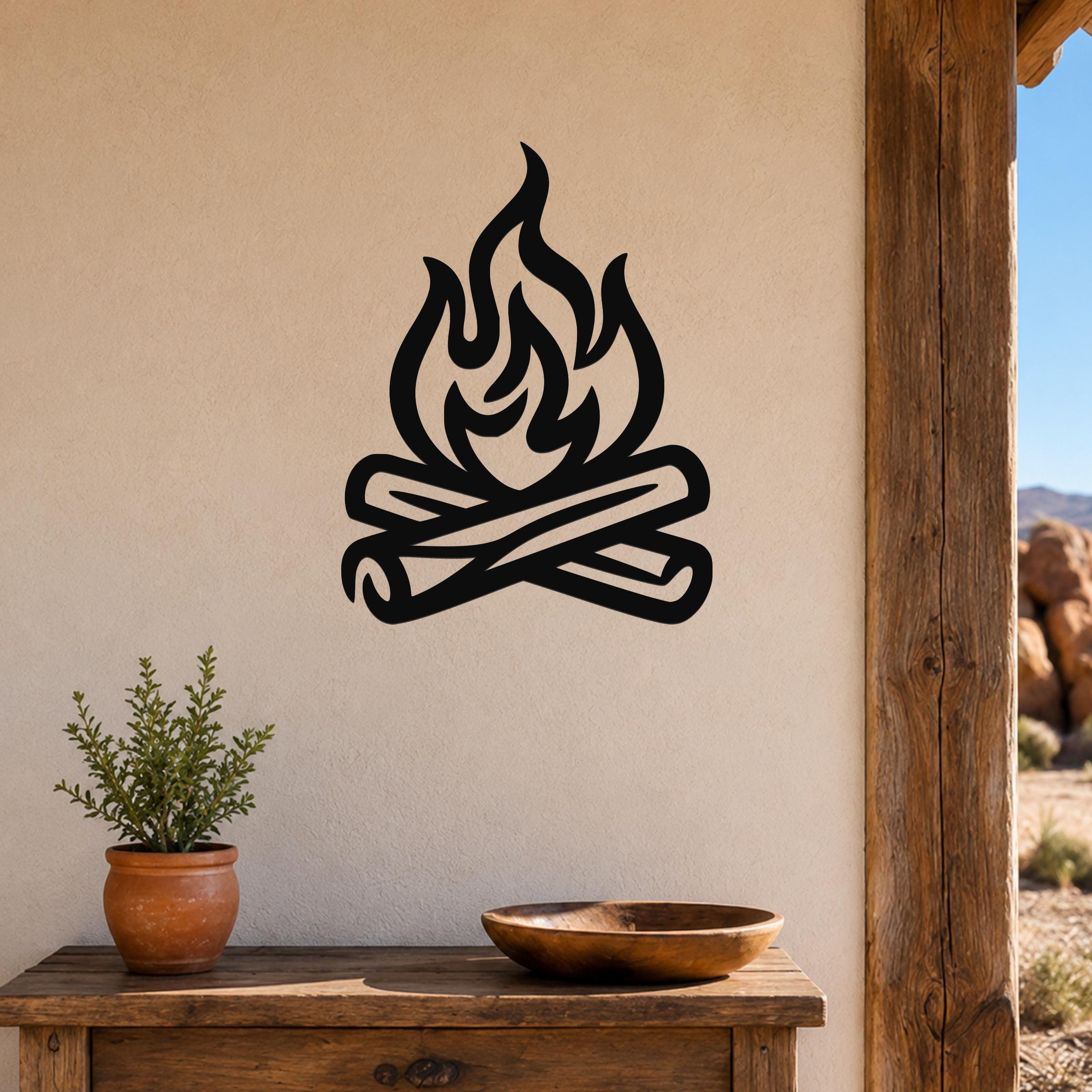 Black metal campfire wall art with flame and log silhouette, mounted on a beige wall above a wooden table in a rustic outdoor patio setting.