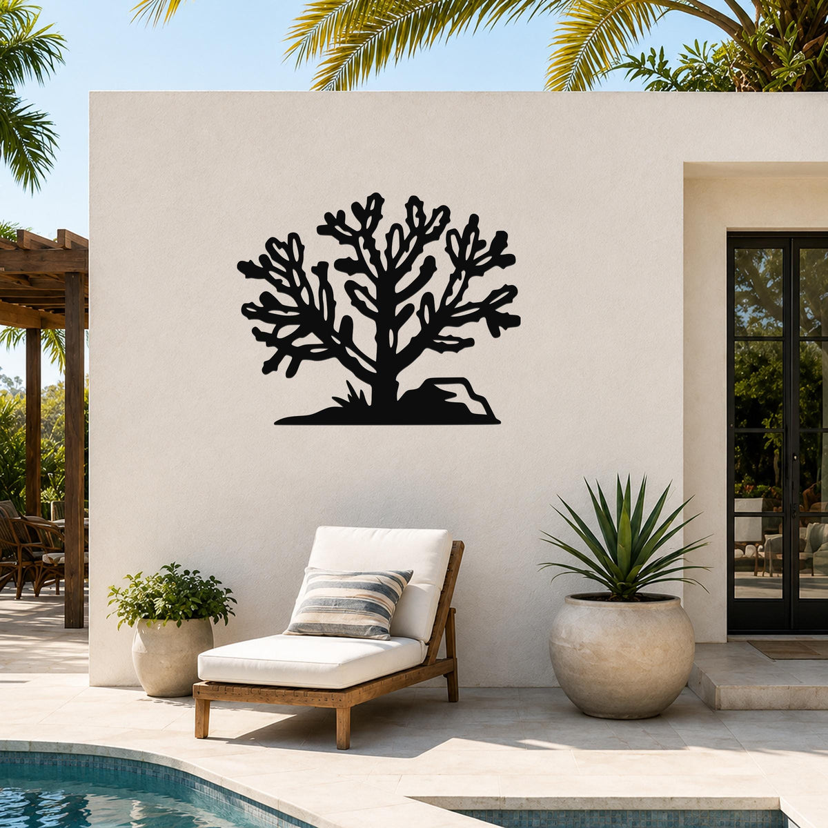 Black metal Cholla cactus wall art with branching cutouts, mounted on a white outdoor wall above a lounge chair near a pool.