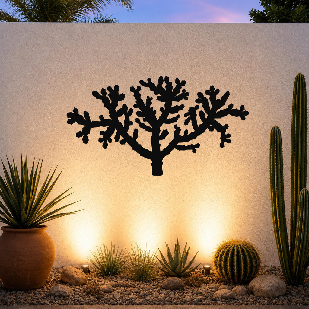 Black metal Cholla cactus wall art with branching cutout silhouette, mounted on a light-colored outdoor wall above a desert garden with potted succulents and ground lights.