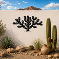Black metal Cholla cactus wall art with spiky branch cutouts, mounted on a white outdoor wall above a desert garden with real cacti and rocks.