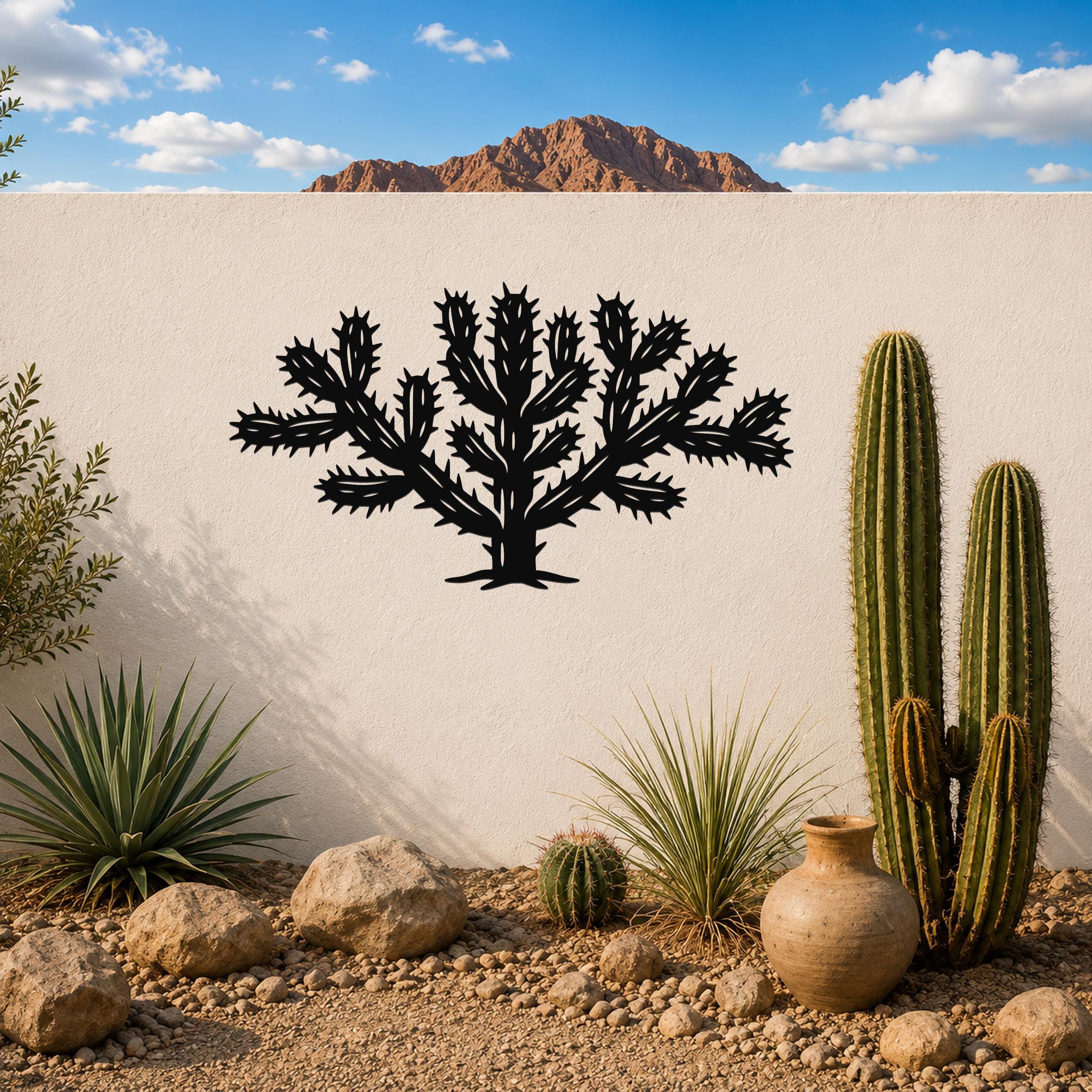 Black metal Cholla cactus wall art with spiky branch cutouts, mounted on a white outdoor wall above a desert garden with real cacti and rocks.
