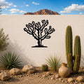 Black metal Cholla cactus wall art with detailed branch cutouts, mounted on a white outdoor wall above a desert garden with real cacti and rocks.
