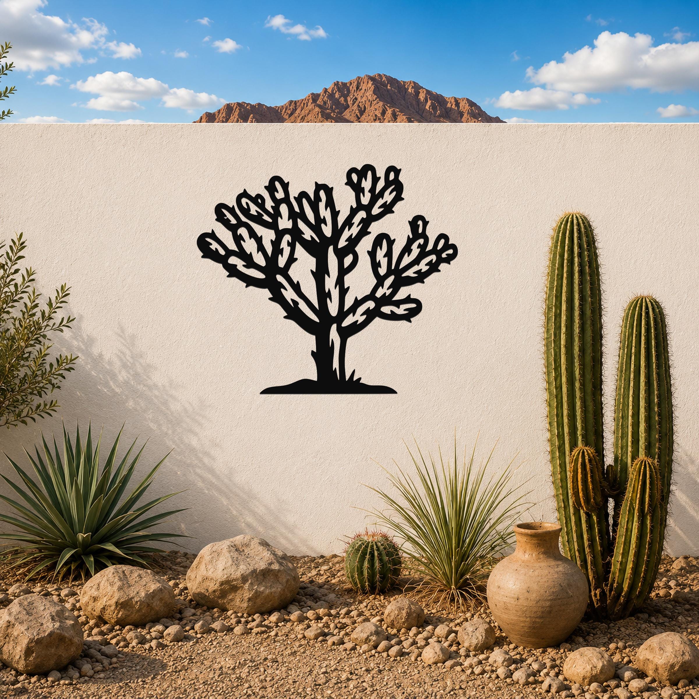 Black metal Cholla cactus wall art with detailed branch cutouts, mounted on a white outdoor wall above a desert garden with real cacti and rocks.