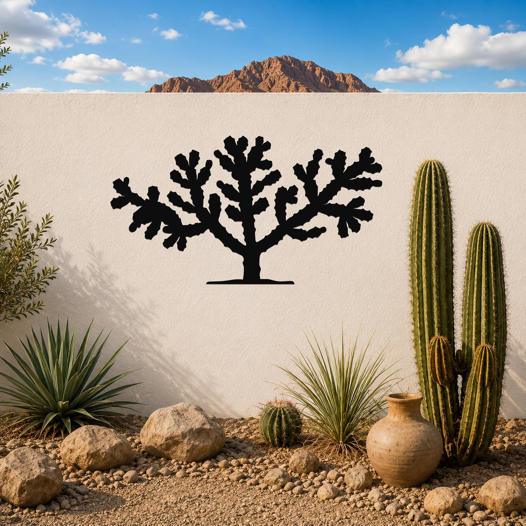 Black metal Cholla cactus wall art silhouette mounted on a white outdoor wall above a desert garden with cacti and rocks.