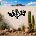 Black metal Cholla cactus wall art silhouette mounted on a white outdoor wall above a desert garden with cacti and rocks.